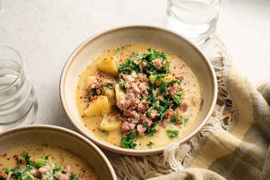 A bowl of creamy Copycat Olive Garden Zuppa Toscana with potatoes, sausage, kale, and visible seasonings sits beside a glass of water and a striped cloth.
