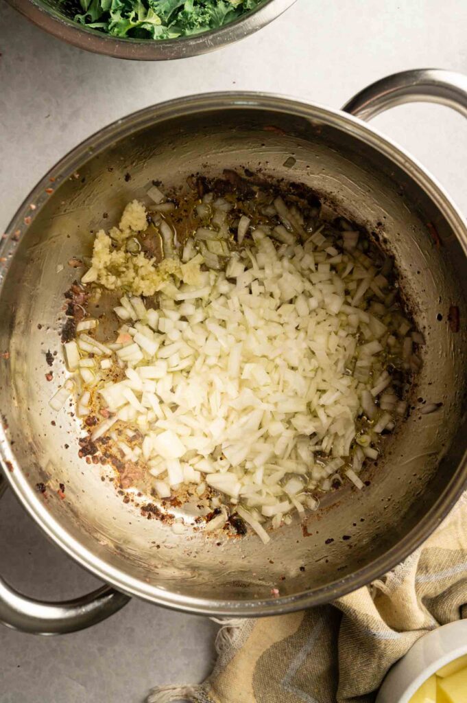 Chopped onions and minced garlic sautéing in oil in a stainless steel pot—a savory base for Copycat Olive Garden Zuppa Toscana, with a towel and bowl partially visible beside it.