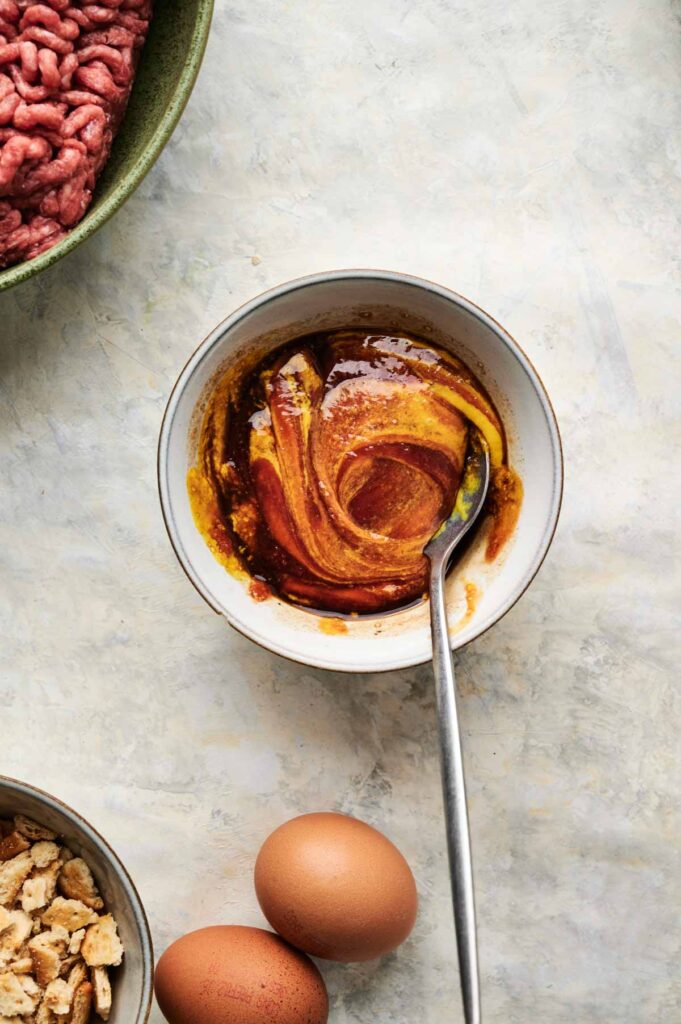 A bowl with a yellow and brown sauce mixture and a spoon, next to eggs, cracker pieces, and a bowl of raw ground meat—perfect ingredients for making Air Fryer Cracker Barrel Meatloaf.
