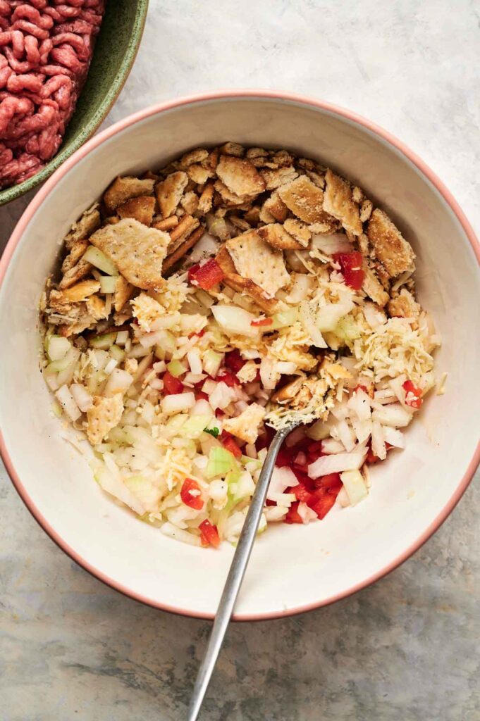 A mixing bowl contains chopped onions, diced red peppers, shredded cheese, and crushed crackers with a spoon—classic ingredients for Air Fryer Cracker Barrel Meatloaf. A bowl of raw ground meat is partially visible nearby.