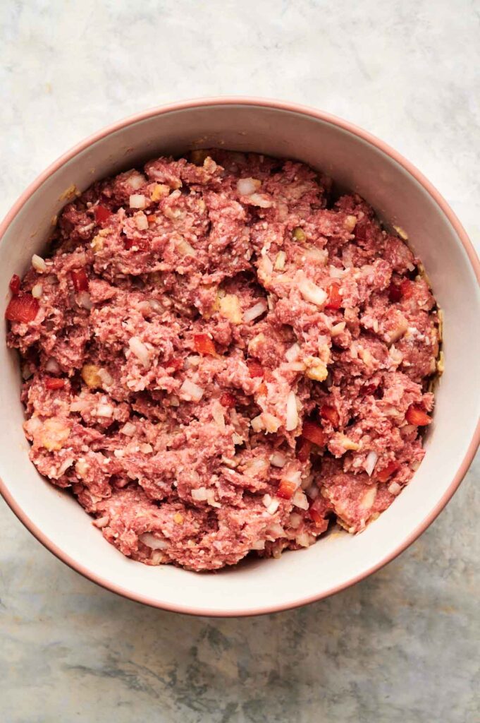 A bowl filled with a mixture for Air Fryer Cracker Barrel Meatloaf, featuring raw ground meat, chopped onions, and diced tomatoes on a light-colored surface.
