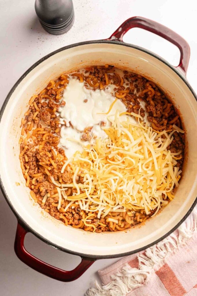 A pot with cooked ground meat, macaroni, shredded cheese, and a creamy white sauce being mixed together on a countertop, creating a delicious Hamburger Helper Cheeseburger Macaroni-inspired meal.