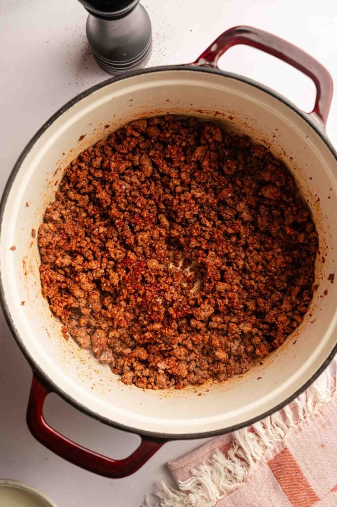 Ground beef cooked with spices for a delicious Hamburger Helper Cheeseburger Macaroni, served in a red and white pot on a light surface beside a striped cloth and pepper grinder.