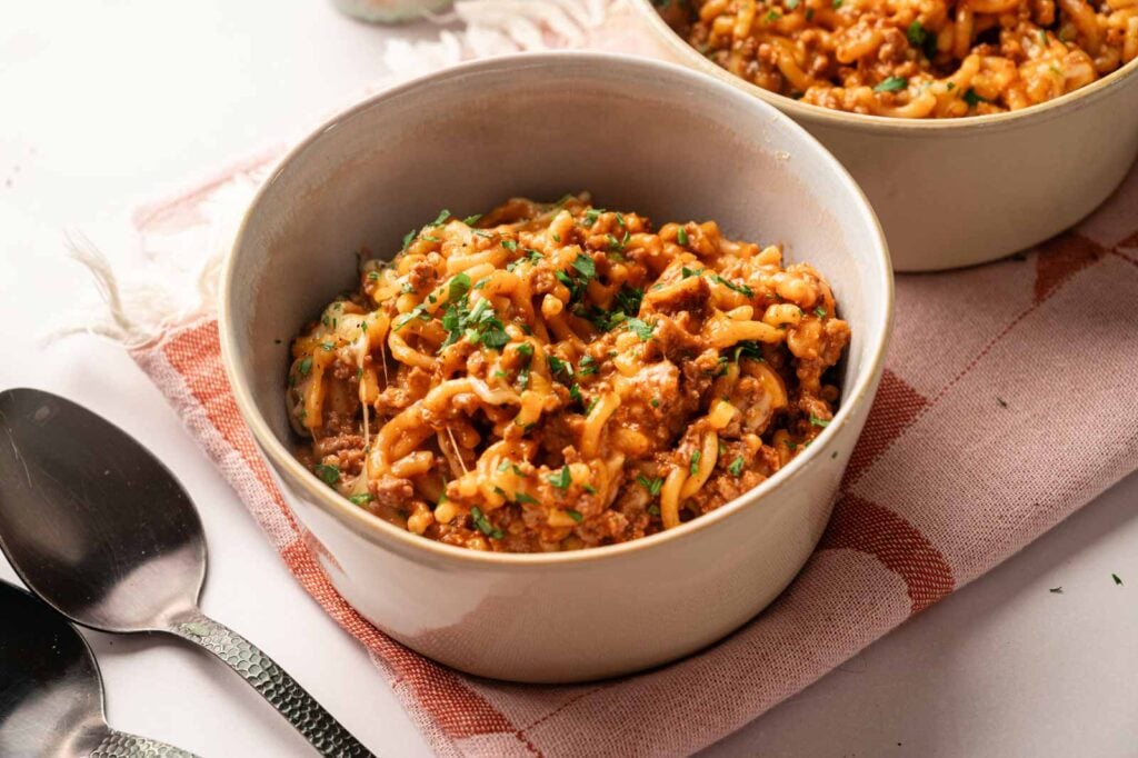 A bowl of Hamburger Helper Cheeseburger Macaroni is topped with chopped herbs and placed on a pink checkered cloth next to two spoons.