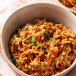 A bowl of cheesy ground beef pasta, inspired by Hamburger Helper Cheeseburger Macaroni, topped with chopped parsley and served on a light-colored cloth with a fork nearby.