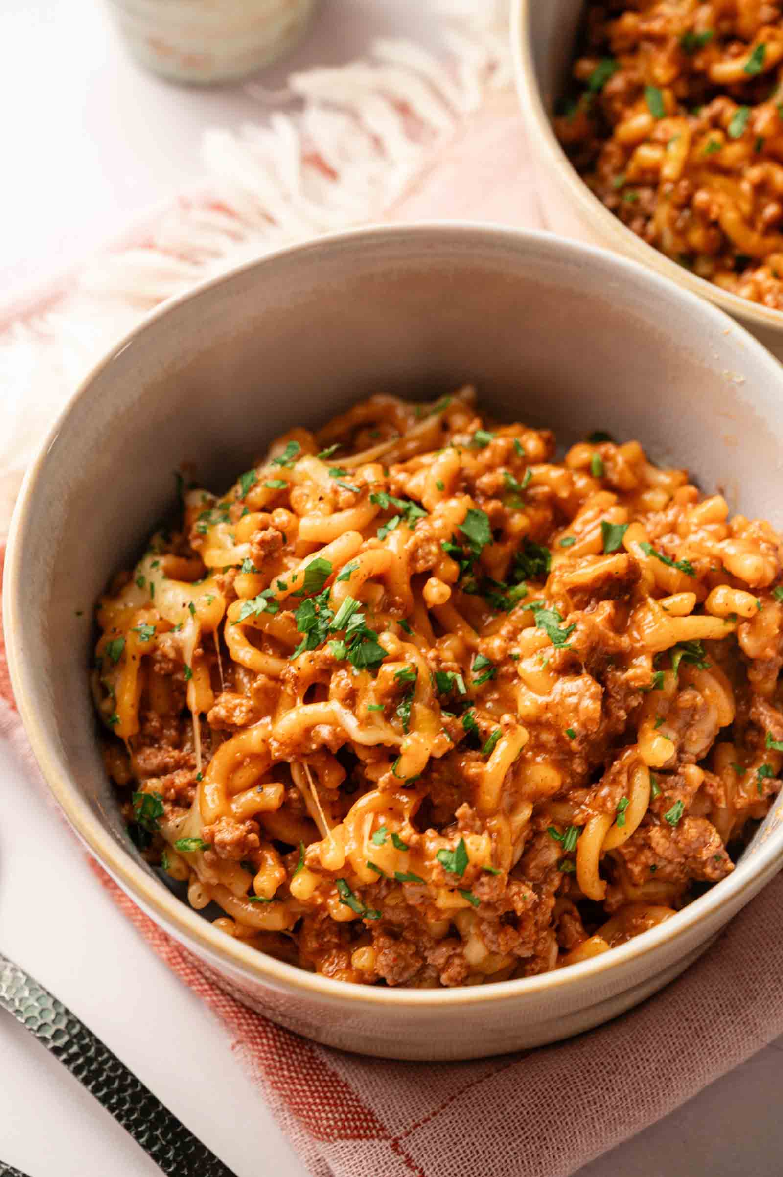 A bowl of cheesy ground beef pasta, inspired by Hamburger Helper Cheeseburger Macaroni, topped with chopped parsley and served on a light-colored cloth with a fork nearby.