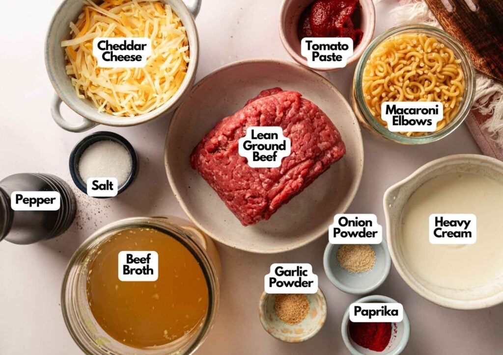 A top-down view of ingredients for a classic Hamburger Helper Cheeseburger Macaroni dish, featuring lean ground beef, cheddar cheese, tomato paste, macaroni elbows, heavy cream, beef broth, and various seasonings.
