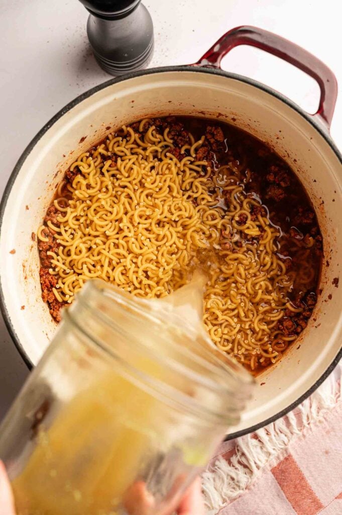 A jar pours broth into a pot containing dry noodles and a red sauce with ground meat, just like the beginning steps for making Hamburger Helper Cheeseburger Macaroni.