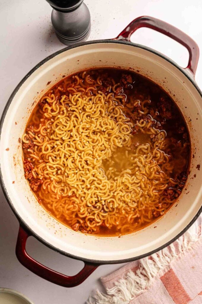 A pot of cooked pasta with the seasoned beef sits on a white surface next to a pink and white fringed cloth, reminiscent of the comforting look of Hamburger Helper Cheeseburger Macaroni.