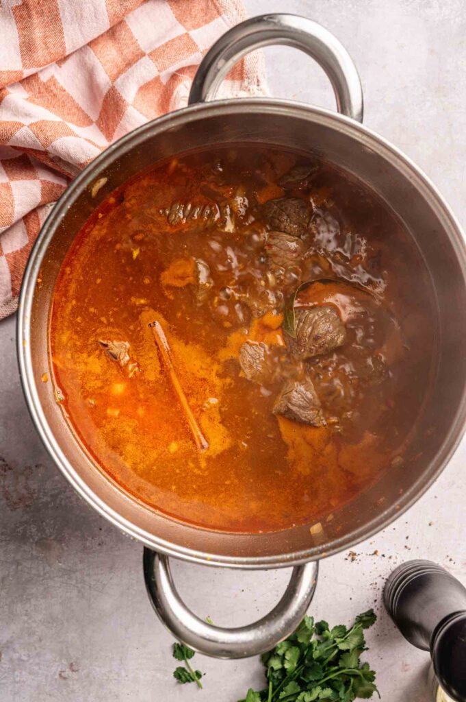 A stainless steel pot filled with simmering beef stew, visible chunks of meat, herbs, and a reddish broth rests on a light countertop beside a checkered towel—perfect for enjoying an Air Fryer Lamb Rogan Josh.
