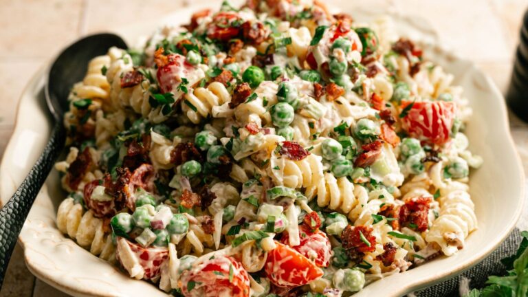 A bowl of rotini pasta salad with peas, cherry tomatoes, bacon bits, chopped herbs, and a creamy dressing, served with a black spoon.