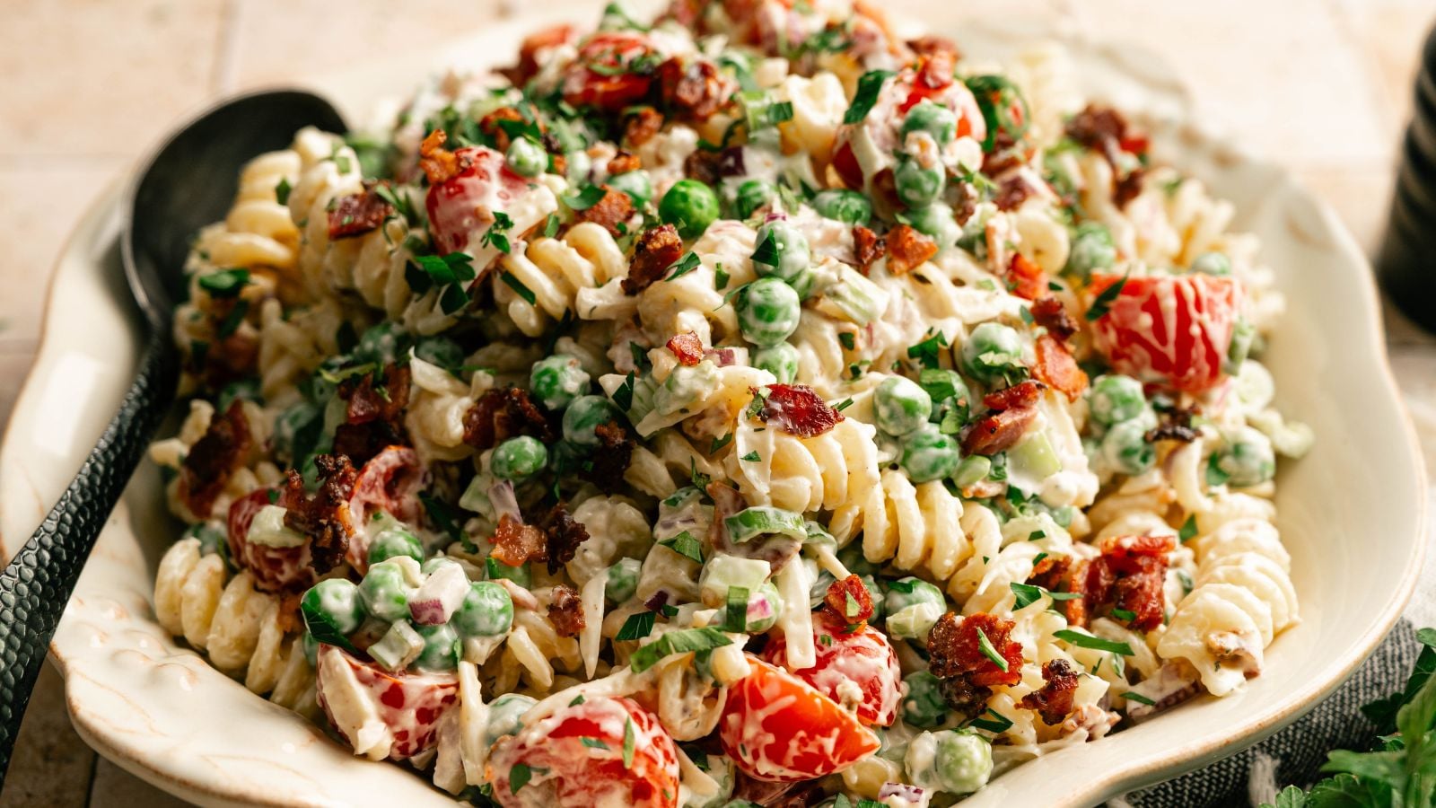 A bowl of rotini pasta salad mixed with peas, cherry tomatoes, bacon bits, chopped herbs, and a creamy dressing, with a black serving spoon beside the dish.