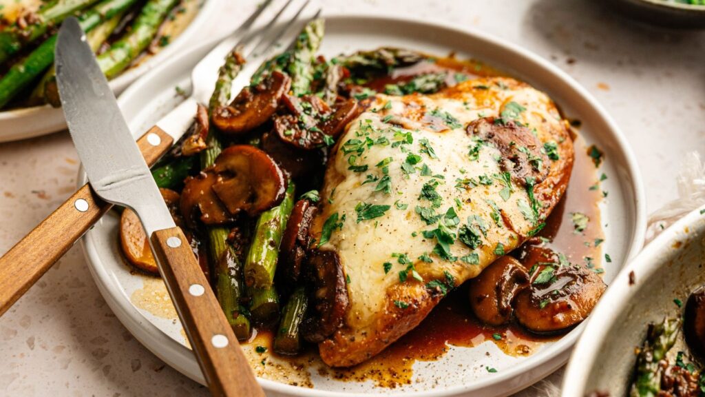 A plate of chicken breast topped with melted cheese and herbs, served with sautéed mushrooms and asparagus, with a knife and fork on the side.