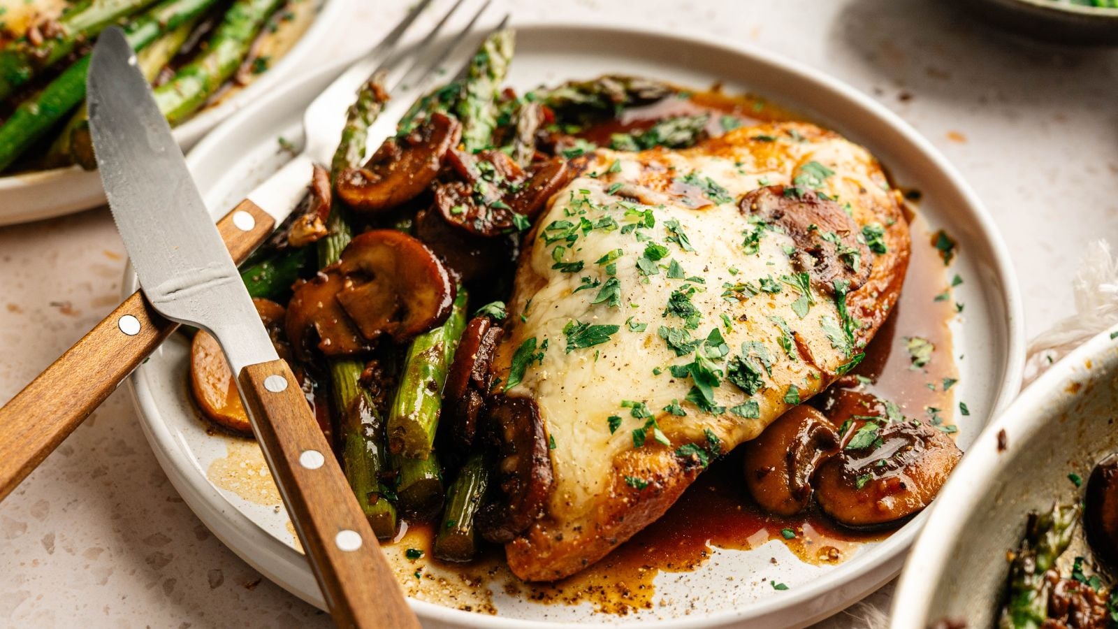 A plate of chicken breast topped with melted cheese and herbs, served with saut&eacute;ed mushrooms and asparagus, with a knife and fork on the side.