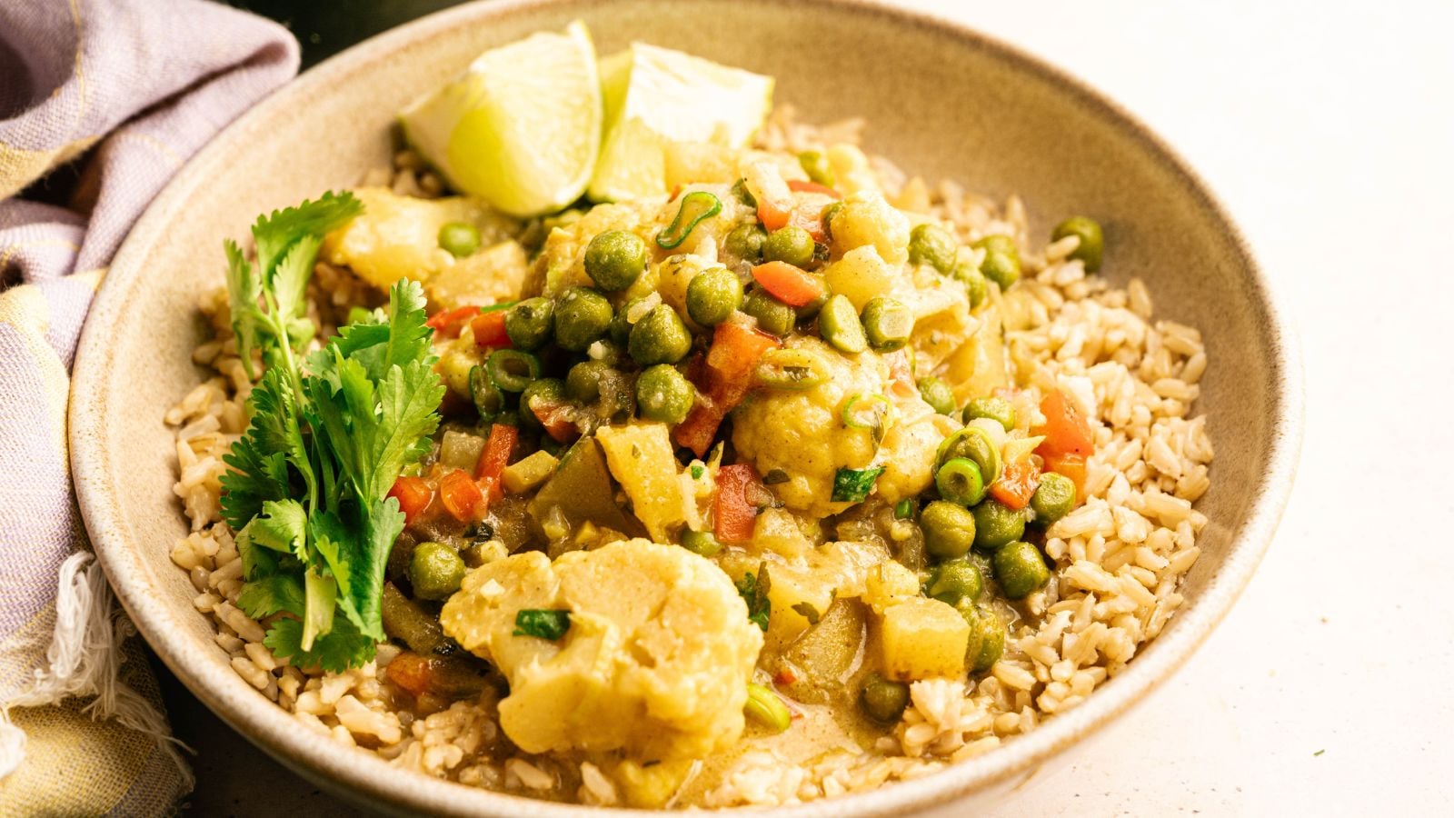 A bowl of brown rice topped with mixed vegetable curry, including peas, cauliflower, and carrots, garnished with fresh cilantro and lime wedges.