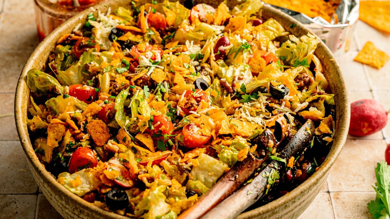 A large bowl of taco salad with lettuce, cherry tomatoes, shredded cheese, olives, ground beef, and crushed tortilla chips, with salad tongs resting on the edge.