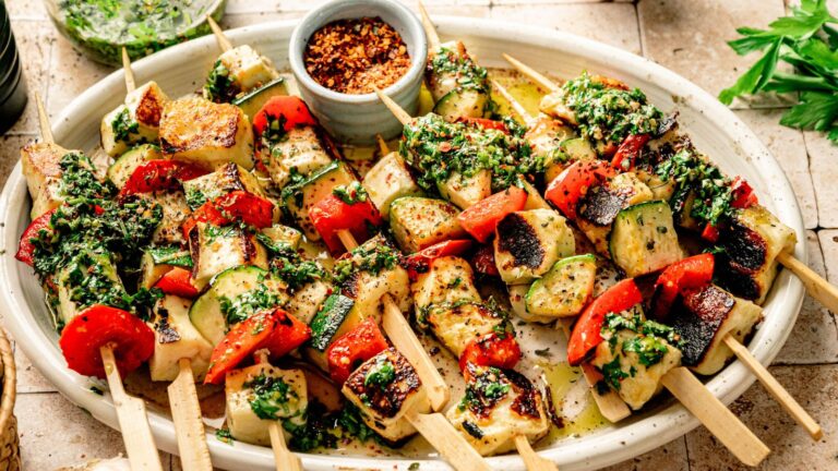 A platter of grilled vegetable and chicken skewers garnished with green herb sauce, with a small bowl of red pepper flakes in the center.