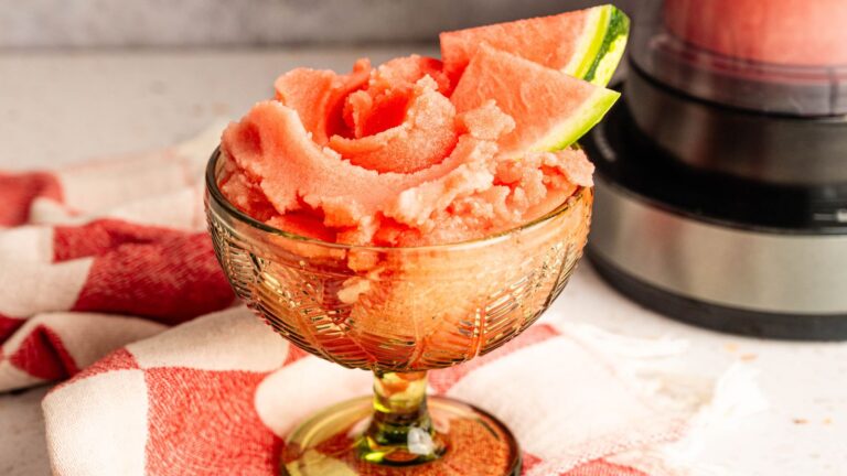 A glass dish filled with watermelon sorbet, garnished with a slice of fresh watermelon, sits on a red and white cloth next to a blender.