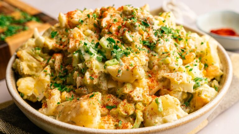 A bowl of potato salad topped with chopped parsley and a sprinkle of paprika, with visible chunks of potatoes, celery, and hard-boiled eggs.