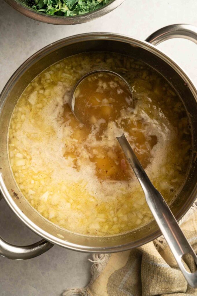 A ladle rests in a pot filled with simmering Copycat Olive Garden Zuppa Toscana broth and chopped onions.